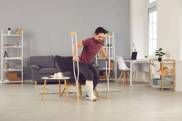 Man with broken leg trying to stand up from chair and walk with crutches in the living-room — Fotografia de Stock