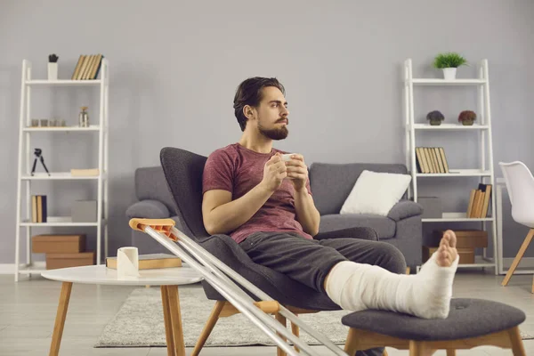 Man with broken leg sitting in armchair, thinking about his injury and feeling bored and lonely — ストック写真