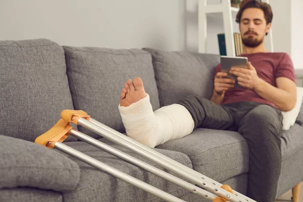 Young man sitting on sofa with broken leg in cast — ストック写真