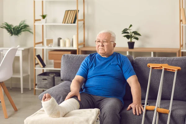 Disappointed senior mature man sitting on sofa with broken leg in cast — Stock fotografie