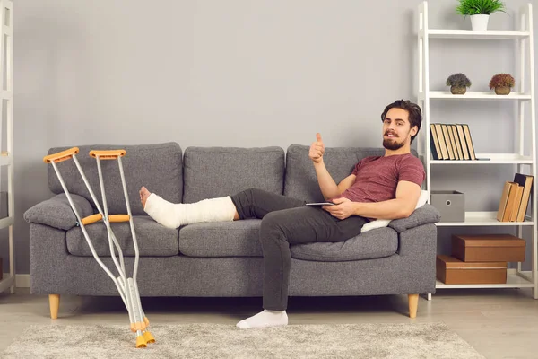 Happy man with broken leg in cast sitting on sofa, using tablet and giving thumbs-up — 스톡 사진