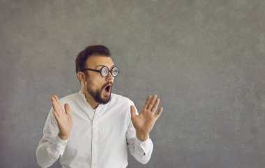 Portrait of funny nerd man in glasses with surprised facial expression on gray background.