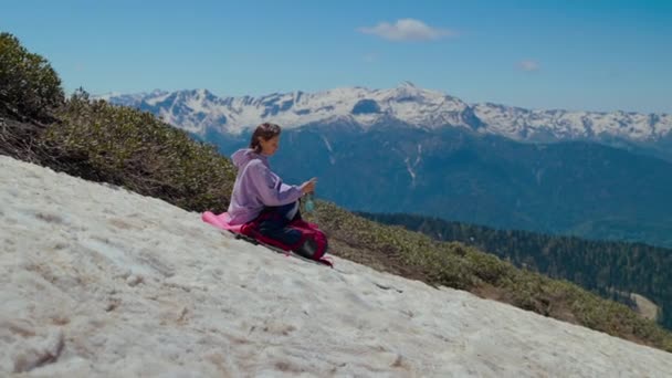 Deteniéndose en la ladera del monte durante el senderismo, la joven está bebiendo agua y admirando las vistas — Vídeos de Stock