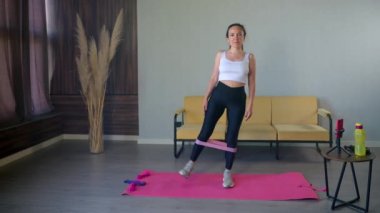 Genç ve güzel bir kadın evde tek başına antrenman yapıyor. Lastik bantla egzersiz yapıyor.