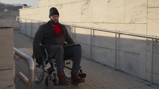 Дорослий інвалідний чоловік в ручному інвалідному візку рухається на рампі в сучасному місті — стокове відео