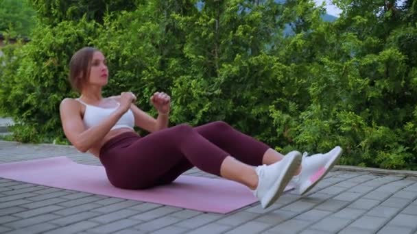 Active woman is training in park in summer day, lifting torso lying on ground and moving arms — Video