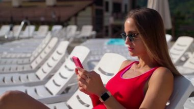 Genç seksi kadın yazın tatil köyündeki modern akıllı telefondan sosyal medyaya bakıyor.