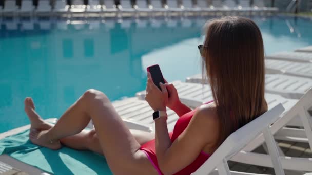 Νεαρή γυναίκα μαυρίζει κοντά σε εξωτερική πισίνα, ξαπλωμένη σε lounger και χρησιμοποιώντας smartphone — Αρχείο Βίντεο