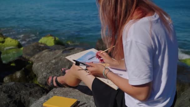 Молодая женщина рисует морской пейзаж, сидя на камне на берегу, творческое хобби — стоковое видео