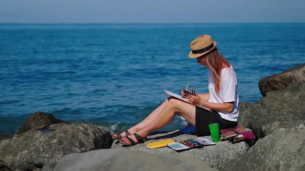 Женщина-художник рисует пейзаж сидя на скалистом берегу моря — стоковое видео