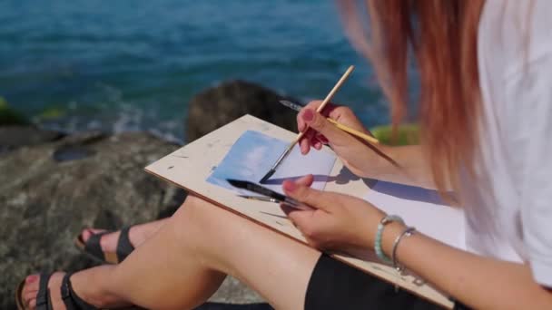 Female student of school of art is painting marine landscape sitting on coast — Stock Video