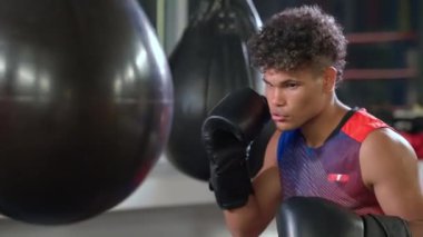 Boks salonunda antrenman yapan Latin bir sporcunun yavaş çekim çekimi. Boksör, agresif ve odaklanmış bir bakışla kum torbasına yumruk atıyor.