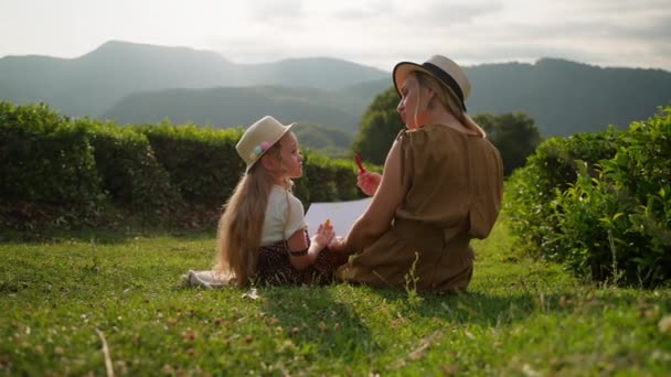 年轻的妈妈带着小女儿坐在绿地的草地上 妇女教孩子从大自然中画画 山景景致 — 图库视频影像