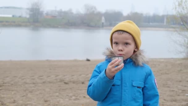 Un niño bebe té en un día frío en el fondo de un río de la ciudad — Vídeo de stock