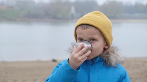 在寒冷的天气里，一个男孩子在城市河流的背景下喝茶 — 图库视频影像