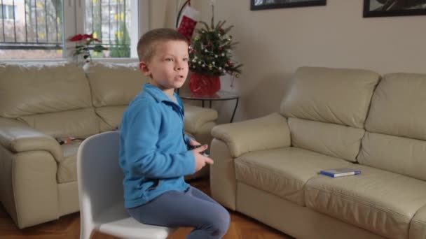 En femårig pojke spelar en spelkonsol nära en julgran. — Stockvideo