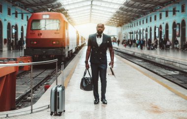 Zarif, kel, sakallı bir Afrikalı hippi. Yelek ve papyon takmış şık bir hippi. Tren istasyonunun peronunda duruyor.
