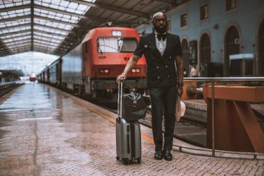 Zarif takım elbiseli, yelekli ve papyonlu, modaya uygun, kel bir Afro-Amerikan girişimci, tren istasyonunda bavullarıyla, kırmızı tren istasyonunun yanındaki platformda duruyor.