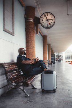 Süslü, kel, siyahi bir girişimci, tren istasyonunun peronundaki ahşap bankta oturuyor, bavullarıyla, ve trenini beklerken saatine göre kontrol ediyor.