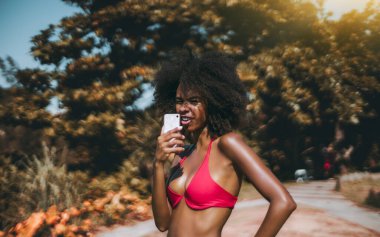 Kırmızı bikinili komik, genç, seksi bir Brezilyalı kadın, güneşli bir günde plajın yanındaki sahil parkında dururken akıllı telefonuyla bir sesli mesaj yazdırarak sohbete cevap veriyor.