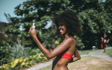 Bikinili komik bir Afro-Amerikalı kadın video kaydediyor ve akıllı telefonuyla yayın yapıyor ya da tropik bir tatil beldesinin plaj bölgesinde dururken bir arkadaşıyla görüntülü görüşme yapıyor.