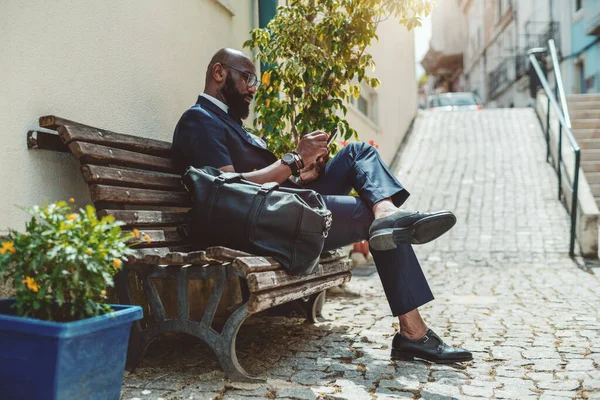 A handsome mature bearded bald black man entrepreneur in an elegant outfit is sitting on a wooden street bench on pavement and using his smartphone; African-American businessman is phoning outdoor