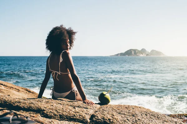 入り江の岩の上に座って 振り返ってみると素晴らしい若い黒人女性 ジャネイロ海岸で日光浴をしながらココナッツ水で海岸に白いビキニを身に着けているかわいい髪の女性 — ストック写真