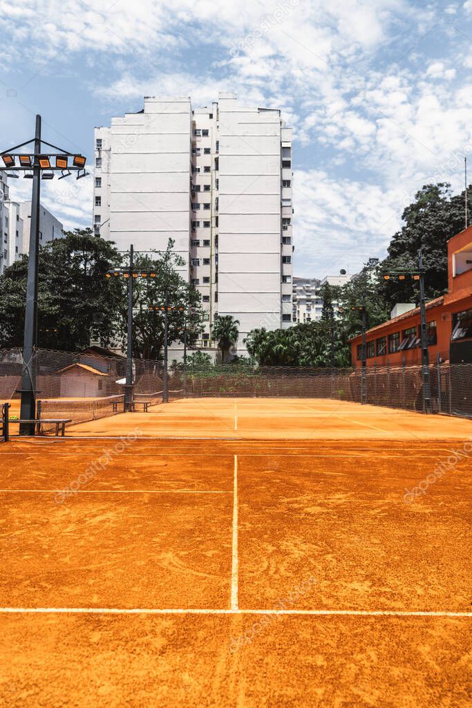 An outdoor view of a sports club with four tennis courts with clay surfaces, safety nets, and spotlight poles. An empty tennis club with residential buildings, trees, and blue sky in the background