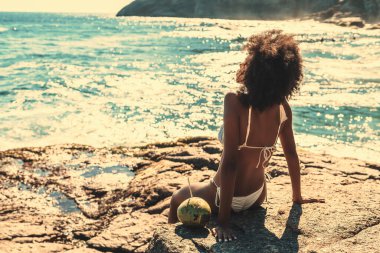 View from behind of a ravishing young African American woman with curly African hair tanning while sitting on the rock of a beach and enjoying dazzling sunlight with a teal oceanand coco next to her clipart