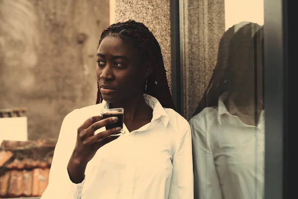 迷人的深色皮肤女人的画像 手里拿着小玻璃杯 凝视着某个东西或某个人 非洲一名辫子女 靠着玻璃门 边喝咖啡边望着旁边 — 图库照片