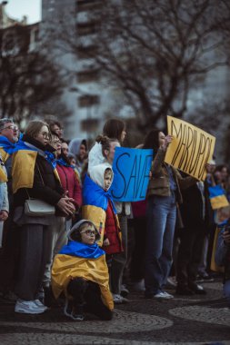 Lisbon, Portugal - April 20, 2022: protest rally dedicated to saving the Ukrainian city of Mariupol from the Russian aggression; a kid in glasses wrapped in flag squats next to other protesters clipart