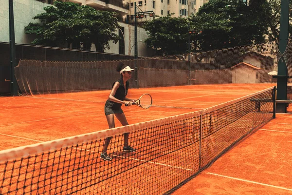 An African-American female tennis player in the practice of receiving position on a court. A young black woman playing sports on a tennis court, standing on orange clay ground on a sunny day