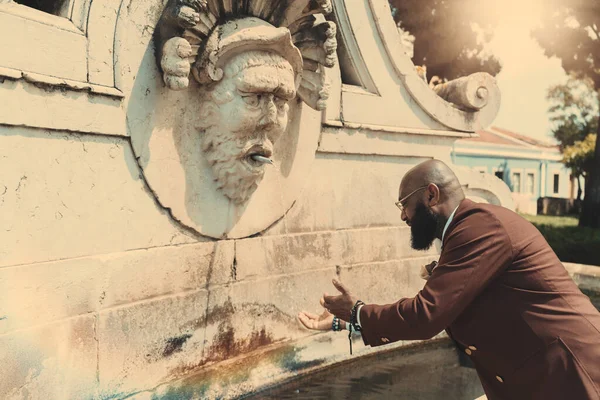 A side view of a mature bald black man with a beard, in a tailored suit of a chestnut color and eyeglasses, washing his hands in the water of a street antique marble fountain or going to drink from it
