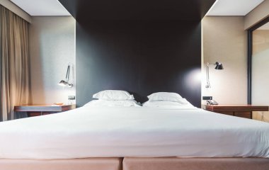 Boş bir otel odasının geniş açılı görüntüsü. Yatağın yan tarafında iki açık gece lambası var. Yastıklara özenli odaklanma.