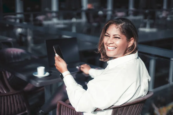 A portrait of a dazzling laughing Hispanic plus-size businesswoman sitting in an outdoor cafe with her laptop and a cup of coffee and holding a smartphone in her hand; a Brazilian woman with cellphone