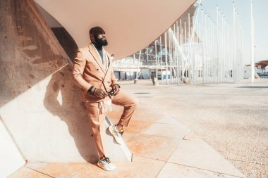 Güzel sakallı, bej rengi kostümlü, beton bir duvara yaslanmış şık bir Afrikalı girişimci, sokakta takım elbise giymiş süslü, kel bir iş adamı.