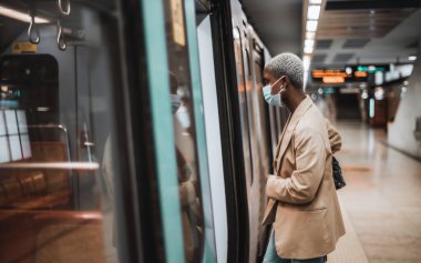 Sığ bir alan derinliği ve kısa saçlı, anti-virüs maskeli, terminaldeki metro istasyonundaki platformdan boş bir metro trenine binen siyahi bir kadına odaklanmış bir görünüm.