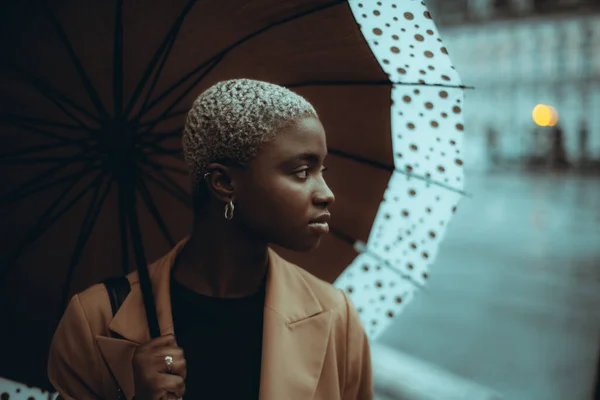 一幅低调的肖像画 描绘了一个心事重重的 可爱的年轻黑人女性 她的短发涂成了白色 站在雨中的街道上 带着一把斑斑的雨伞 她向旁边看了看 右边是一个复制的空间 — 图库照片