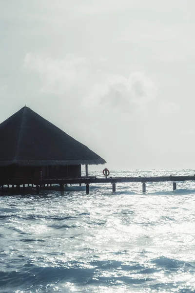 在温暖明亮的天气里 一个有着三角形屋顶的豪华马尔代夫度假胜地的天篷平房和一个带有救生圈的木制码头的垂直拍摄的海景 上面有波涛汹涌的亮点 — 图库照片