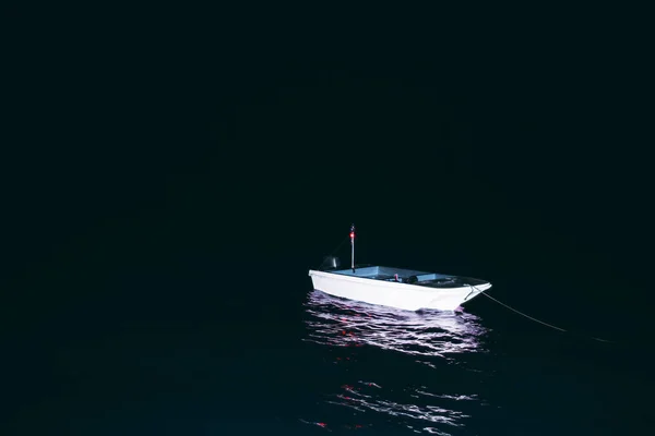 黑暗中的一条孤零零的小船在黑暗中被聚光灯照亮的低调镜头 水面上有红色的灯塔灯和紫色的反光 左边是一个复制的空间区域 — 图库照片