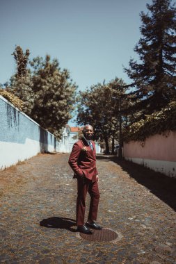 Siyah sakallı, gözlüklü, kestane rengi, kravatlı, caddedeki kaldırım taşının üzerinde duran zarif, olgun, kel bir Afrikalı son sınıf öğrencisinin dikey fotoğrafı.