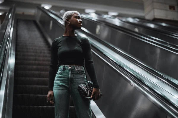 在自动扶梯上的一个年轻非洲姑娘下降到地铁站的低调画像 在通往地铁站或地下通道的移动楼梯上的年轻黑人女子的画像 — 图库照片