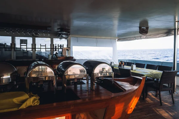 A wide-angle view of an empty ship restaurant with a buffet area, a bar, and one of the tables in the background; a cafeteria zone of a sailing luxury diving safari yacht with the ocean horizon behind