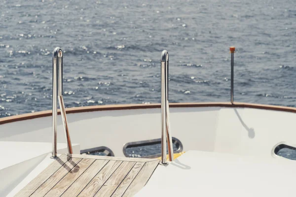 View from a yacht stern of a part of the bow of the boat with a selective focus on a chromed ladder railing and a wooden floor; a beacon light and a blue seascape in a defocused background