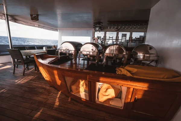 A wide-angle view of an empty ship restaurant interior with an ocean in the background; a safari yacht cafeteria on a middle deck; a cafe on a vessel with a wooden floor, bar counter, and no people