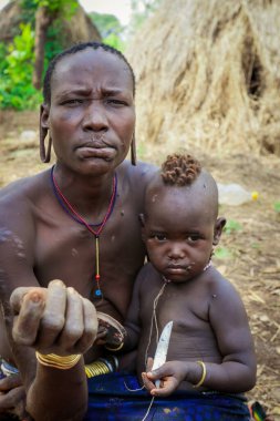 Omo Nehri Vadisi, Etiyopya - 29 Kasım 2020: Kulağında koca delikleri olan bebeği olan kadın Mursi kabilesi köyündeki turistlere poz veriyor