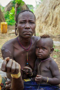 Omo Nehri Vadisi, Etiyopya - 29 Kasım 2020: Kulağında koca delikleri olan bebeği olan kadın Mursi kabilesi köyündeki turistlere poz veriyor