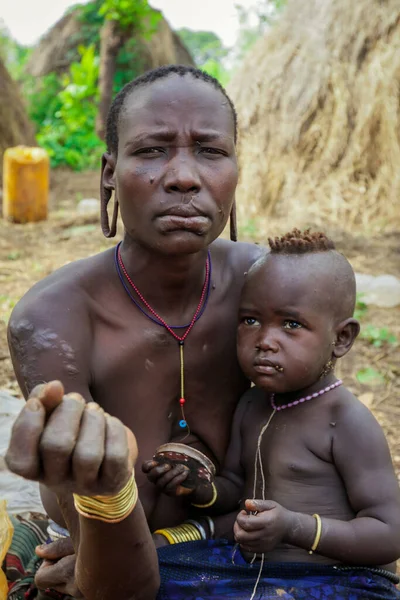 Omo Nehri Vadisi, Etiyopya - 29 Kasım 2020: Kulağında koca delikleri olan bebeği olan kadın Mursi kabilesi köyündeki turistlere poz veriyor