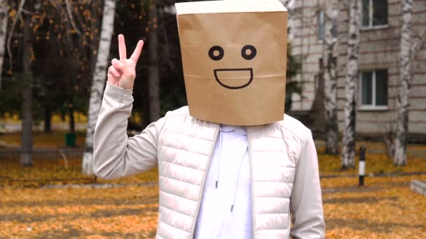 Усміхнений чоловік в паперовій сумці з щасливими емоціями, людина в масці стоїть на відкритому повітрі і показує жест перемоги — стокове відео