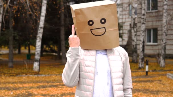 Усміхнений чоловік в паперовій сумці з щасливими емоціями, людина в масці стоїть на відкритому повітрі і демонструє агресивний жест — стокове відео